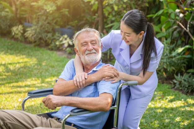 A smiling older man in a wheelchair sits outdoors while a cheerful caregiver in a light uniform leans over and gently hugs his shoulders in a garden setting.
