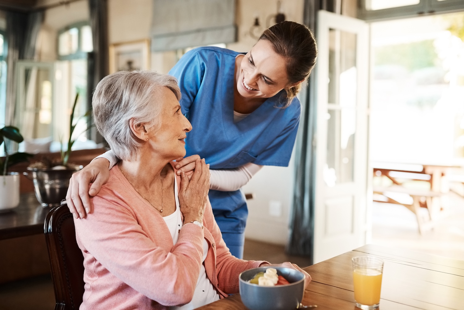 What Are the Eligibility Requirements for Assisted Living A Guide to the Essentials Elderly woman sitting at a dining table with a bowl of fruit and a glass of juice, smiling warmly at a caregiver in blue scrubs who stands beside her with a supportive hand on her shoulder.